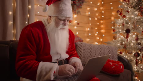 Ein-Porträt-Eines-Echten-Weihnachtsmanns,-Der-An-Einem-Laptop-Arbeitet-Und-Am-Heiligabend-Zu-Hause-Auf-Der-Couch-Spazieren-Geht.-Der-Weihnachtsmann-Beantwortet-E-Mails,-Durchsucht-Die-Internetbank-Und-Arbeitet-An-Einem-Laptop